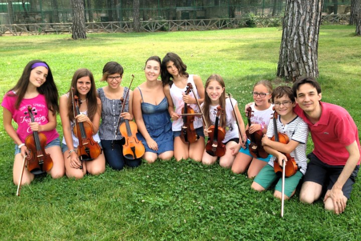 Grupo de alumnos de violín junto a sus profesores durante una actividad musical al aire libre.
